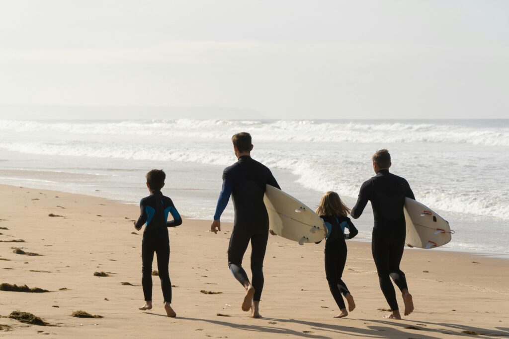 surfing cornwall. a family runs along the shoreline in wetsuits holding surfboards.